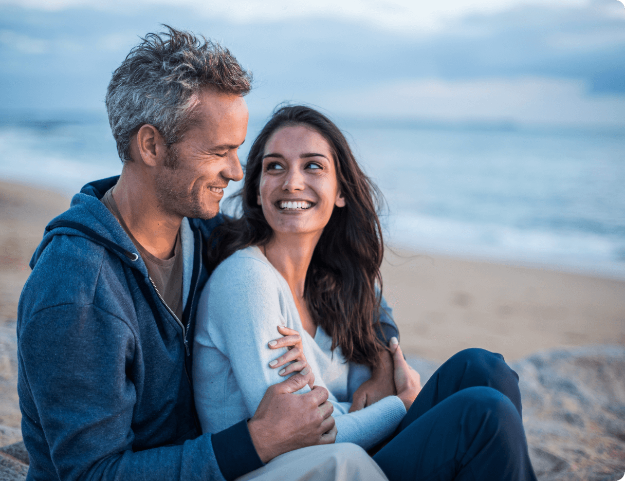 Male and female couple sitting close together on beach with ocean in background