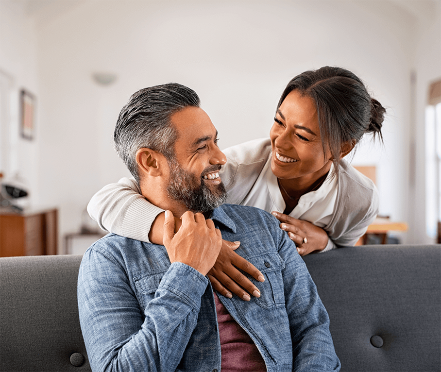Man and woman couple near couch smiling at each other with woman's arm around the man.